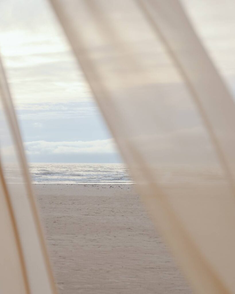Blick durch Vorhänge auf Strand und Meer bei sanftem Licht